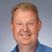 Der Mann hat eine freundliche Ausstrahlung, mit einem breiten Lächeln und hellblauen Augen vor einem grauen Hintergrund.