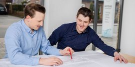 Ein Mann zeigt mit einem roten Stift auf einen Plan, während der andere aufmerksam zuhört und nachdenklich wirkt.