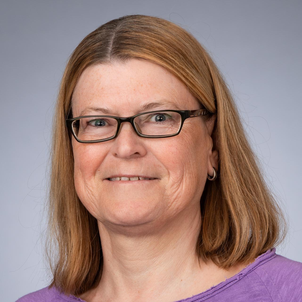 Mit einem sanften Lächeln blickt die Frau in die Kamera, ihre Brille und das lila Shirt verleihen ihr einen freundlichen Look.