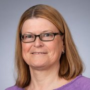 Mit einem sanften Lächeln blickt die Frau in die Kamera, ihre Brille und das lila Shirt verleihen ihr einen freundlichen Look.