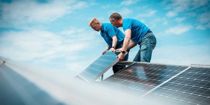 Ein junger Mann und ein älterer Mann heben ein Solarpanel an, um es auf dem Dach zu befestigen, umgeben von Wolken.