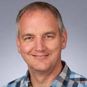 Der Mann hat eine entspannte Ausstrahlung, mit einem breiten Lächeln und blauen Augen, vor einem neutralen Hintergrund.