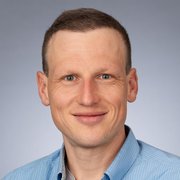 Ein Mann mit kurzen, hellen Haaren und einem freundlichen Lächeln trägt ein blau gestreiftes Hemd vor einem neutralen Hintergrund.