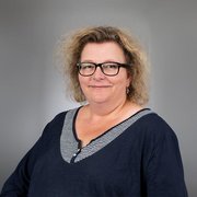 Eine lächelnde Frau mit lockigem, blondem Haar trägt eine dunkelblaue Bluse mit gestreiftem Ausschnitt und steht vor einem grauen Hintergrund.