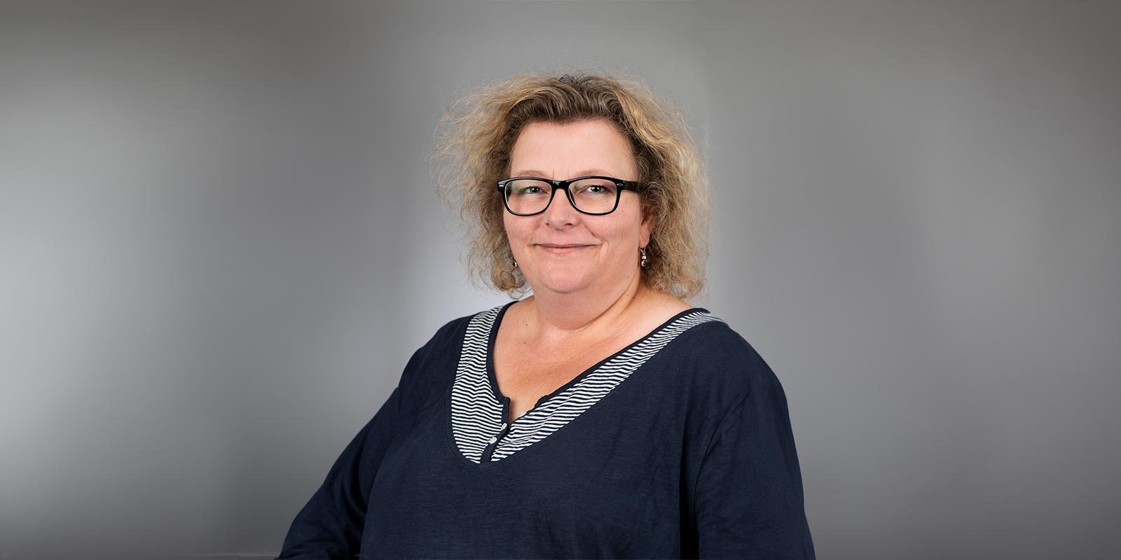 Eine lächelnde Frau mit lockigem, blondem Haar trägt eine dunkelblaue Bluse mit gestreiftem Ausschnitt und steht vor einem grauen Hintergrund.