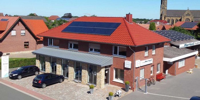 Ein modernes Gebäude mit rotem Ziegeldach und Solarpanelen, umgeben von grüner Landschaft und blauen Autos auf dem Parkplatz.