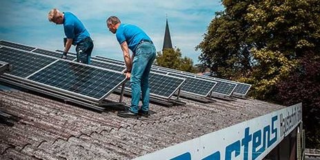 Zwei Arbeiter in blauen T-Shirts heben Solarpanels auf einem Dach, mit einem klaren Himmel und einer Kirche im Hintergrund.