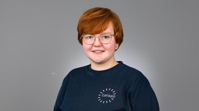 Die Person hat eine positive Ausstrahlung, steht vor einem grauen Hintergrund und trägt ein Sweatshirt mit einem Logo.