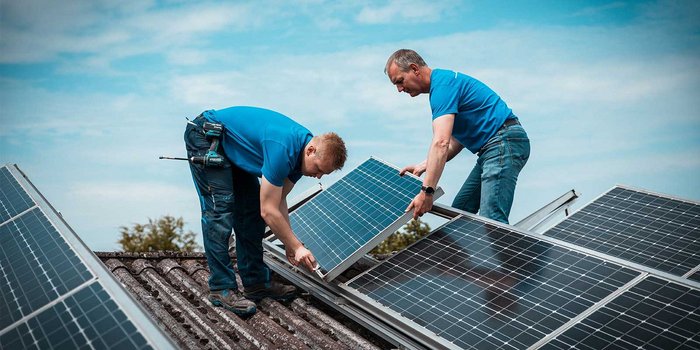 Zwei Männer in blauen T-Shirts befestigen Solarpanels auf einem Dach, konzentriert und engagiert bei ihrer Arbeit.