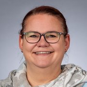 Eine lächelnde Frau mit Brille und einem hellen Schal posiert vor einem neutralen Hintergrund.