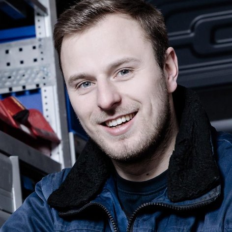 In einer Werkstatt lächelt ein Mann in blauer Arbeitskleidung, während er entspannt auf einem Stuhl sitzt.