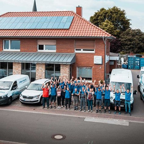 Vor einem modernen Gebäude mit Solarpanelen posiert ein Team in blauen Shirts, umgeben von mehreren Firmenfahrzeugen.