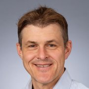 Ein lächelnder Mann mit kurzen, braunen Haaren und einem hellen Hemd vor einem grauen Hintergrund.