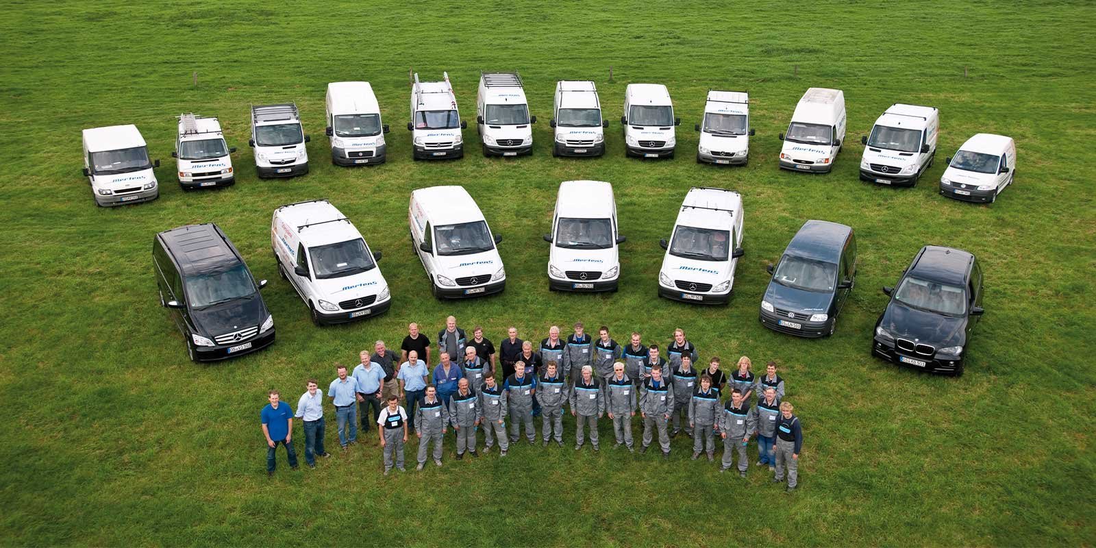 Auf einer grünen Wiese posiert ein Team von Fachleuten vor einer Reihe von weißen und schwarzen Lieferwagen.