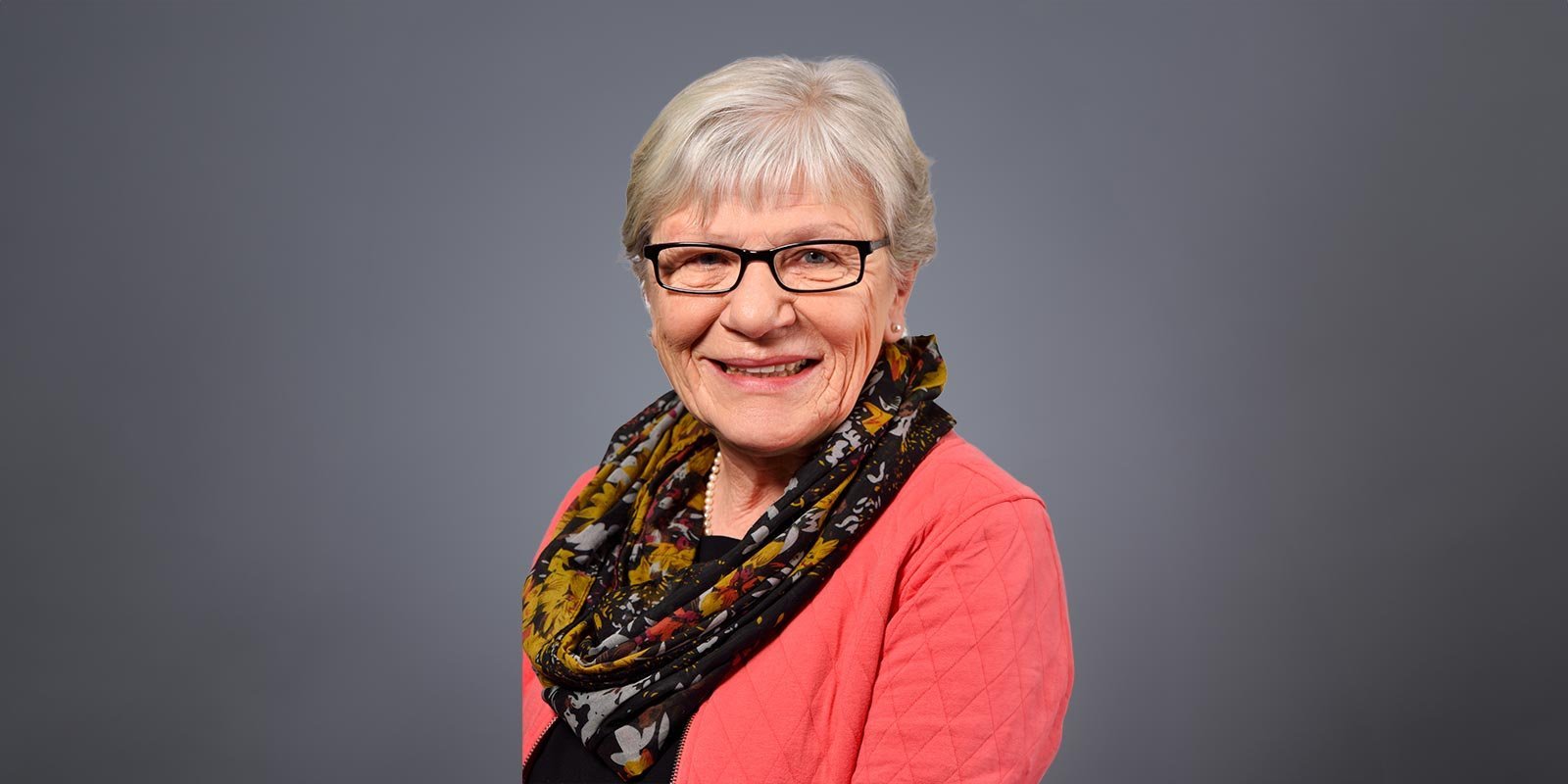 Die Dame mit grauen Haaren und Brille strahlt Lebensfreude aus, während sie in einem eleganten Outfit posiert.