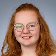 Eine junge Frau mit lockigem, rotem Haar und Brille lächelt freundlich in die Kamera vor einem grauen Hintergrund.