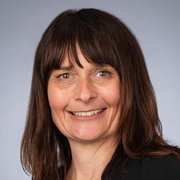 Eine Frau mit langen, braunen Haaren lächelt freundlich in die Kamera, während sie einen schwarzen Blazer trägt.