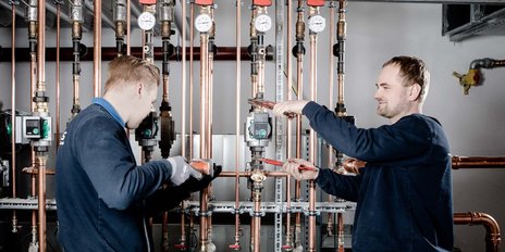 In einem technischen Raum reparieren zwei Männer eine Rohrleitung, während sie Werkzeuge und Messgeräte verwenden.