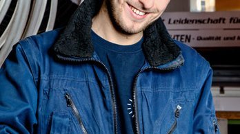 Ein junger Mann in blauer Arbeitsjacke lächelt, während er ein Tablet in einem Lager mit Schläuchen hält.