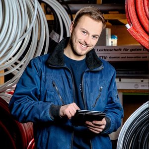 Ein junger Mann in blauer Arbeitsjacke lächelt, während er ein Tablet in einem Lager mit Schläuchen hält.