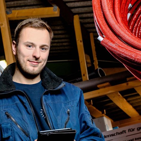 Ein junger Mann in blauer Arbeitskleidung steht in einem Lager mit verschiedenen Kabelrollen und lächelt freundlich.