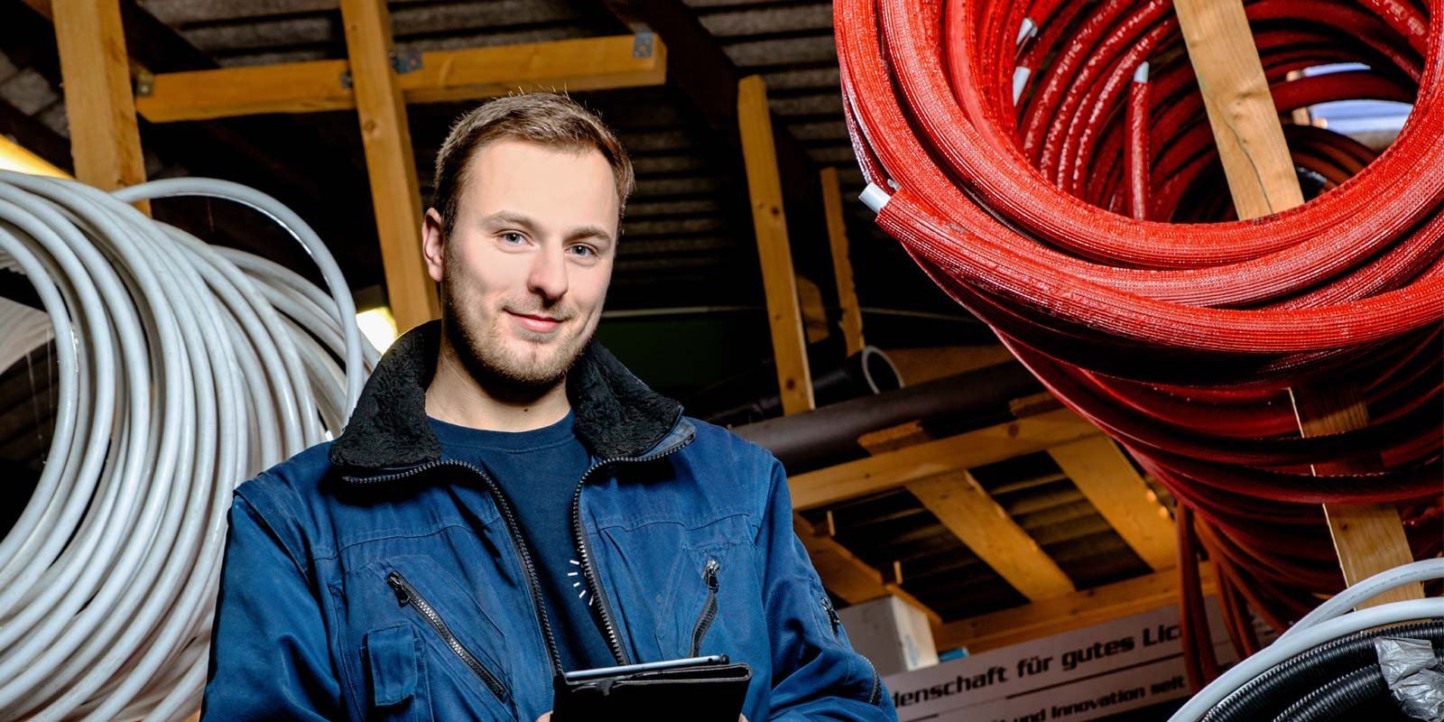 Ein junger Mann in blauer Arbeitskleidung steht in einem Lager mit verschiedenen Kabelrollen und lächelt freundlich.