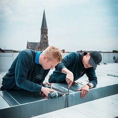 Zwei Handwerker arbeiten konzentriert auf einem Dach, während eine Kirche im Hintergrund sichtbar ist.