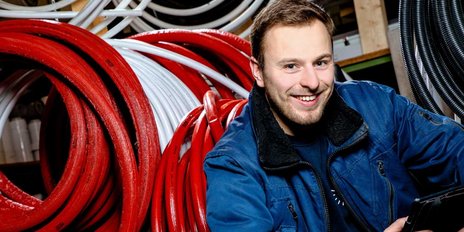 Ein junger Mann lächelt, während er in einem Lager mit bunten Schläuchen aus rotem, weißem und schwarzem Material sitzt.