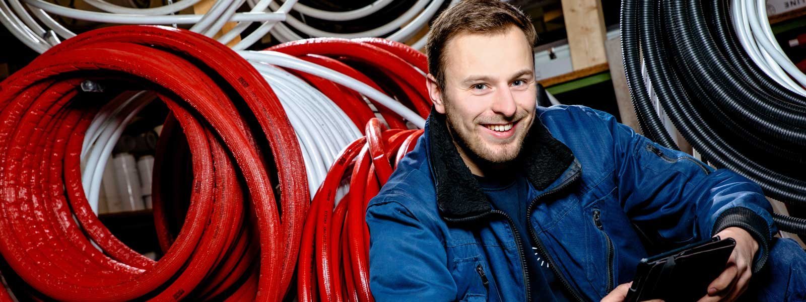 Ein junger Mann lächelt, während er in einem Lager mit bunten Schläuchen aus rotem, weißem und schwarzem Material sitzt.