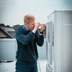 Auf dem Dach eines Gebäudes repariert ein junger Mann eine Klimaanlage, während er sorgfältig ein Werkzeug in der Hand hält.