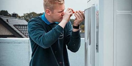 Auf dem Dach eines Gebäudes repariert ein junger Mann eine Klimaanlage, während er sorgfältig ein Werkzeug in der Hand hält.