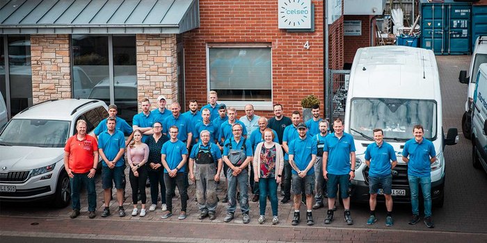 Die Mitarbeiter posieren lächelnd vor ihrem Firmengebäude, umgeben von weißen und blauen Lieferwagen.