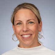 Eine lächelnde Frau mit blonden Haaren trägt große Ohrringe und ein helles Oberteil, vor einem neutralen Hintergrund.