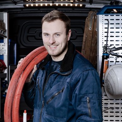 Ein junger Mann in blauer Arbeitskleidung lächelt, während er ein rotes Kabel in der Hand hält, umgeben von Werkzeugen.