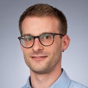 Der Mann hat eine modische Brille und ein Ohrpiercing, sein Gesichtsausdruck ist offen und einladend.