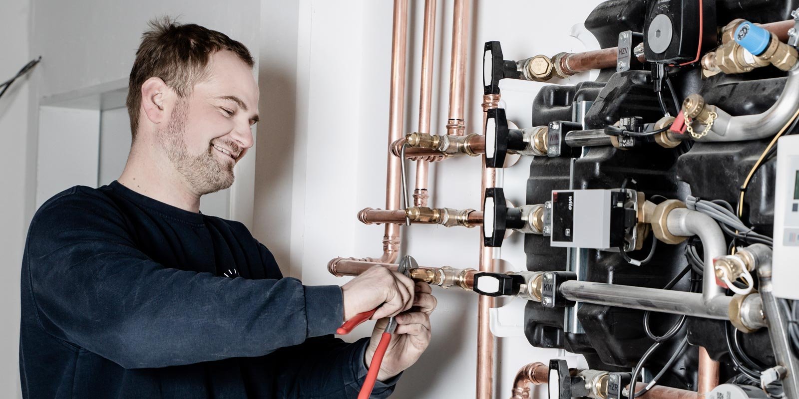 Der Techniker lächelt, während er mit einer Zange an einer Heizungsanlage mit zahlreichen Rohren und Anschlüssen arbeitet.