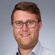 Ein junger Mann mit Brille lächelt freundlich in die Kamera, sein Bart ist gepflegt und er trägt ein kariertes Hemd.