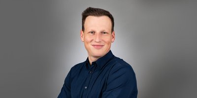 Ein lächelnder Mann mit kurzen, braunen Haaren trägt ein dunkelblaues Hemd und posiert vor einem grauen Hintergrund.