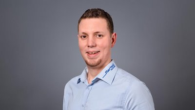 Der Mann hat kurzes, braunes Haar und steht vor einem grauen Hintergrund, während er in die Kamera lächelt.