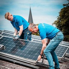 Zwei Männer installieren Solarpanels auf einem Dach, während eine Kirchturmspitze im Hintergrund sichtbar ist.