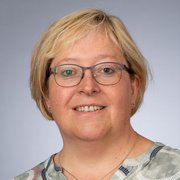 Die Frau trägt ein gemustertes Oberteil und hat eine freundliche Ausstrahlung, während sie direkt in die Kamera schaut.