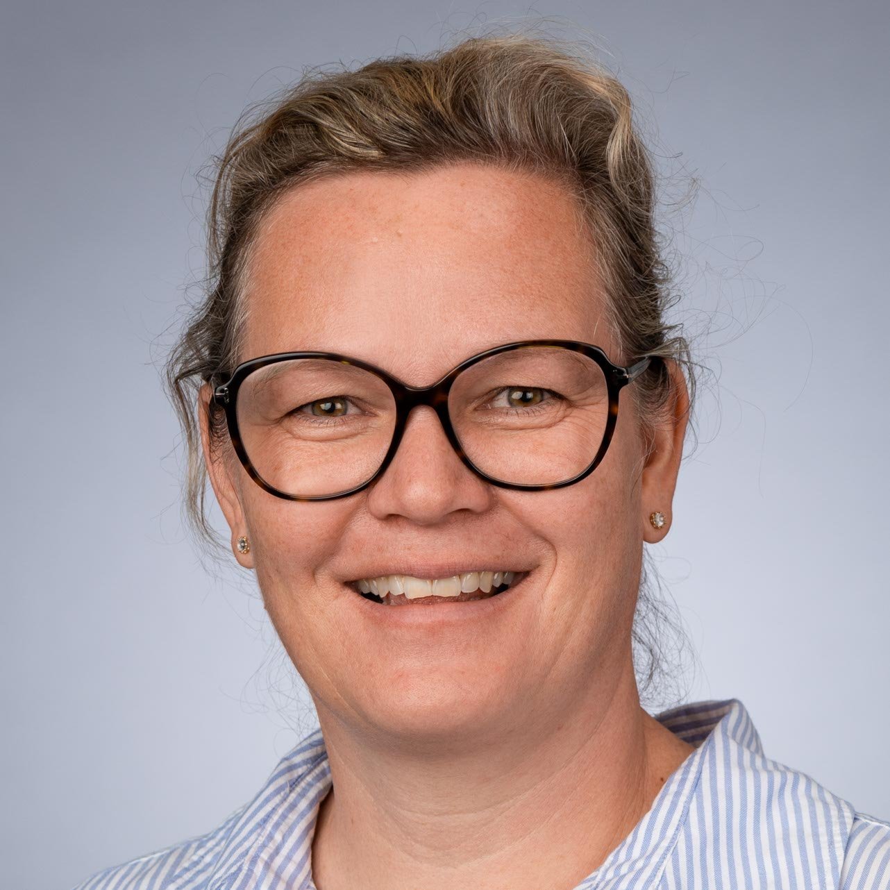 Die Frau hat eine freundliche Ausstrahlung, trägt große Brillen und hat lockige, graue Haare, die lässig fallen.