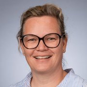 Die Frau hat eine freundliche Ausstrahlung, trägt große Brillen und hat lockige, graue Haare, die lässig fallen.