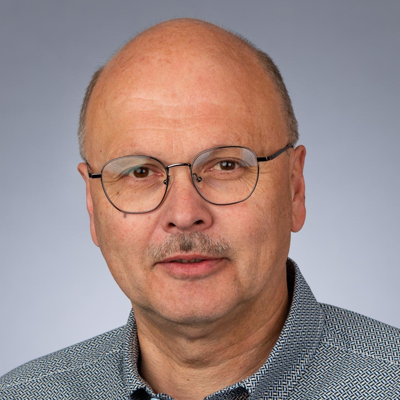 Ein Mann mit Brille und einem leichten Schnurrbart blickt direkt in die Kamera, während er ein gemustertes Hemd trägt.