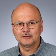 Ein Mann mit Brille und einem leichten Schnurrbart blickt direkt in die Kamera, während er ein gemustertes Hemd trägt.